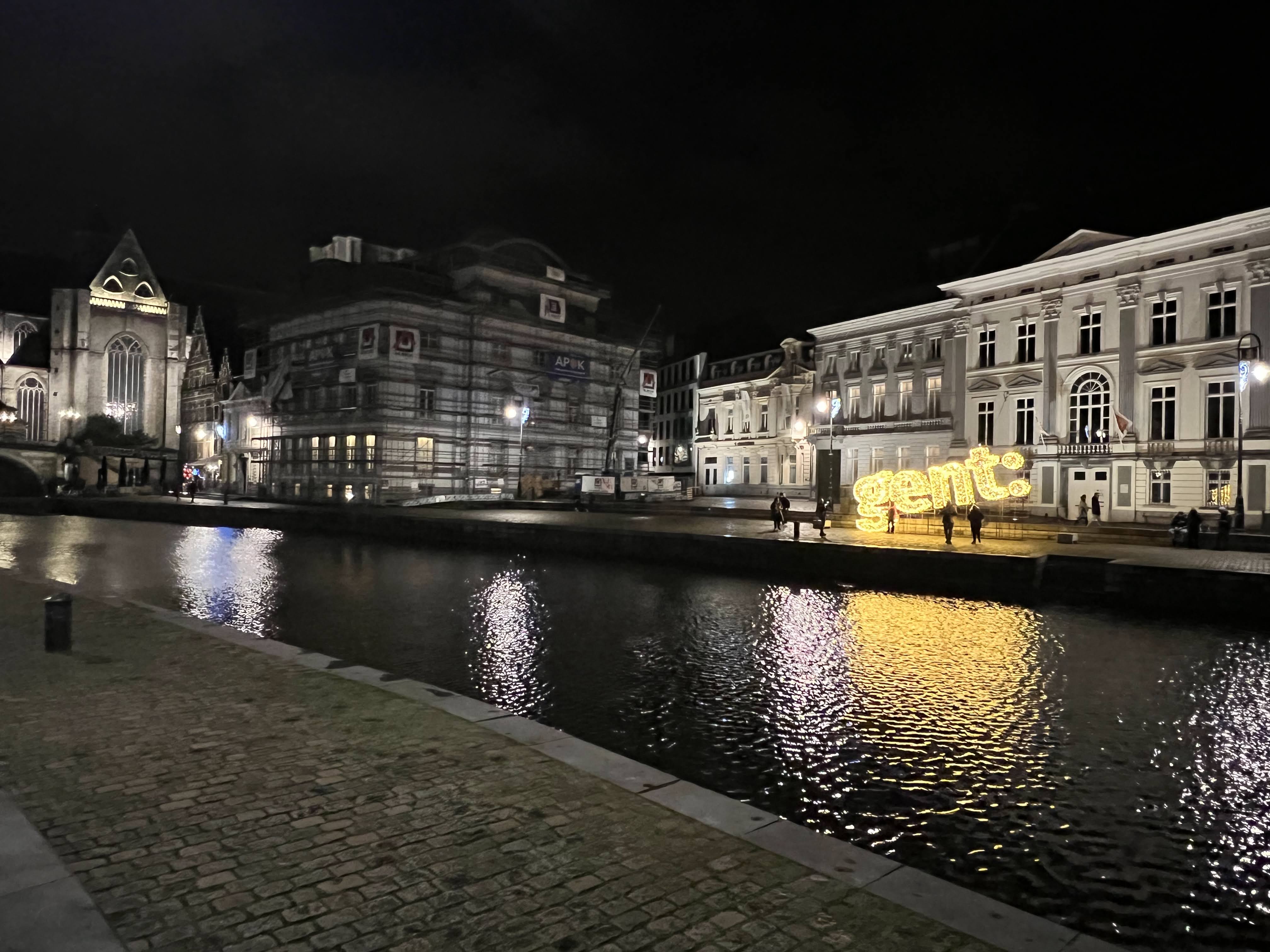 Night time. Stone buildings illuminated with artificial light. Standing along the canal is a sign with illuminated letters spelling Gent: