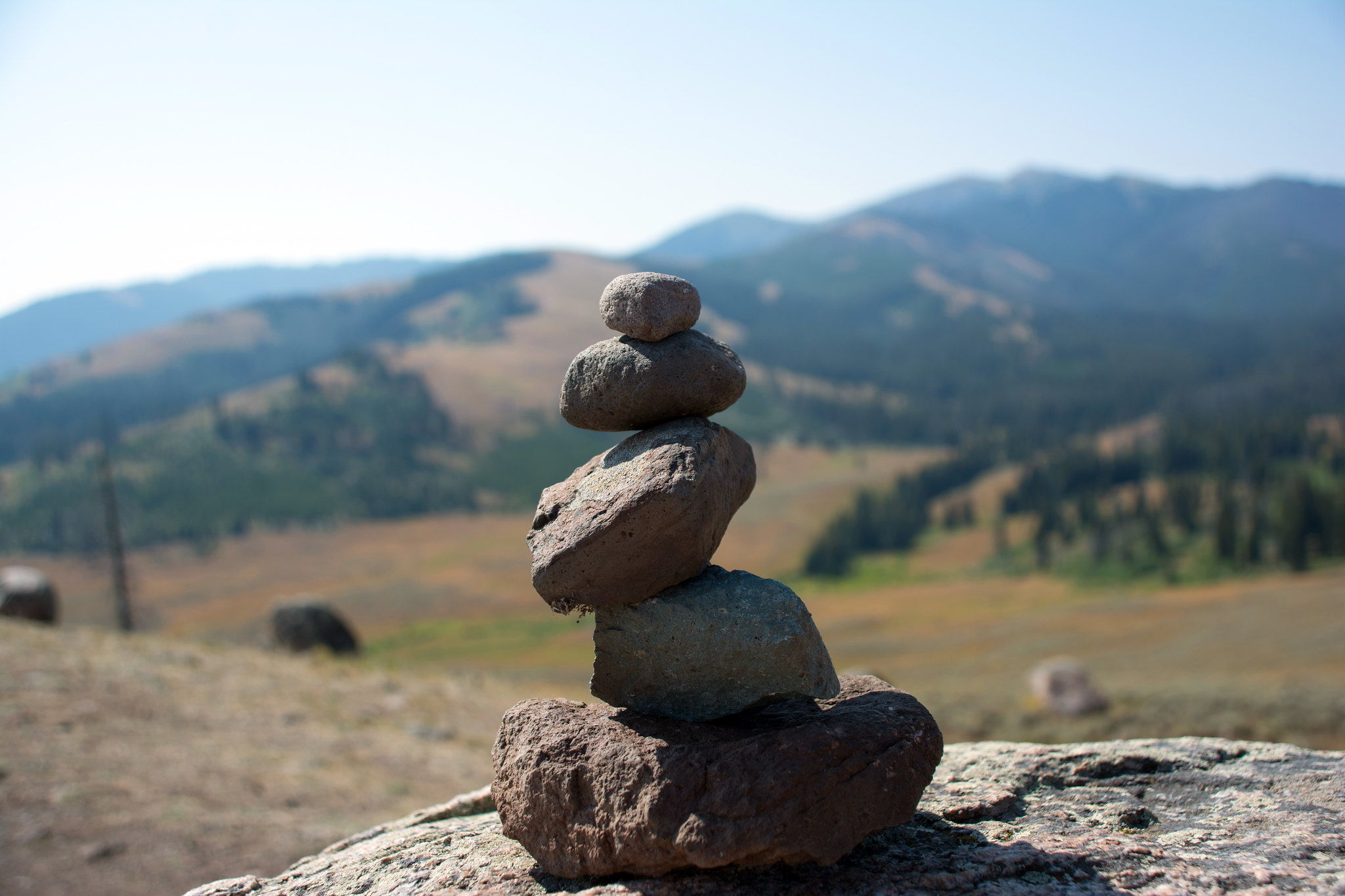 A cairn made of five dark stones stacked vertically upon a large boulder. In the background there are rolling green hills and large green mountains with an overcast sky above.