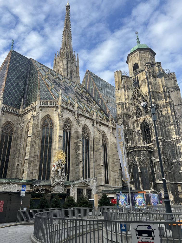 Tall gothic church made of limestone flanked by a blue sky with small white clouds. The roof is adorned with the Austrian coat of arms.
