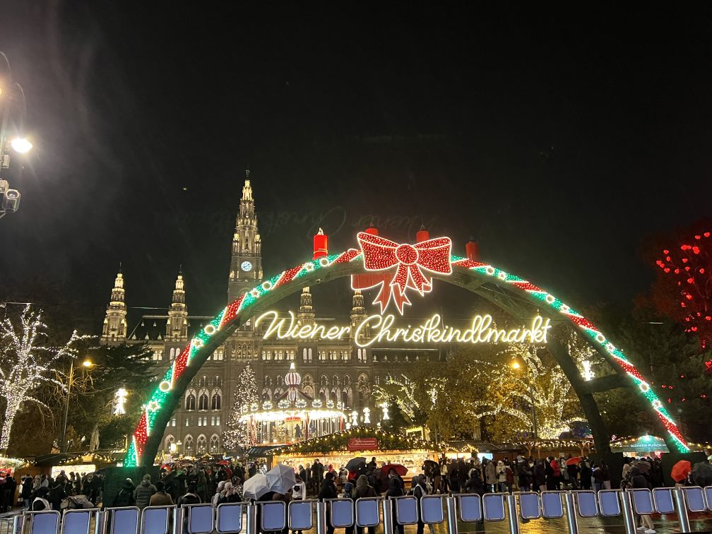 Arch lit with green and red lights and topped with a large red ribbon. It is night time. In the background is the Vienna City Hall and its Christmas Market.