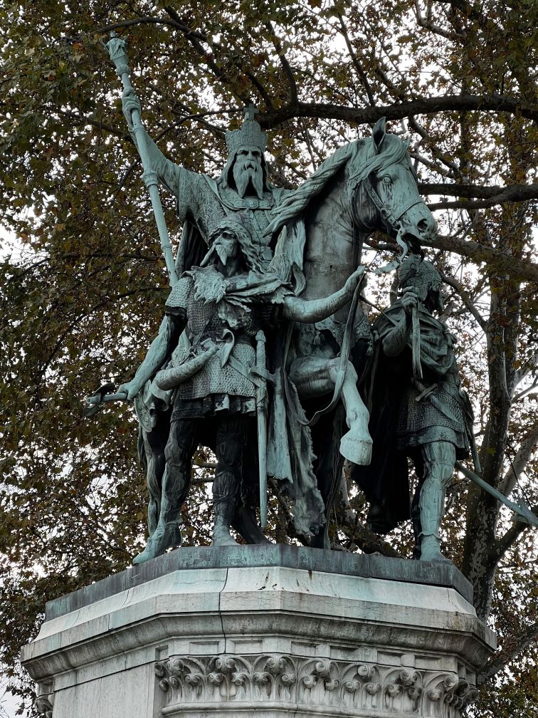 Bronze sculpture of emperor Charlemagne mounted on horseback holding up a staff with his right arm. The statue has a green patina from the weather.