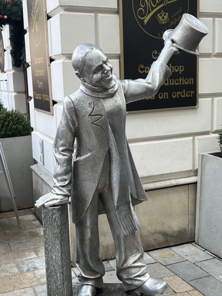 Silver and bronze sculpture of a smiling man. He has removed his tophat with his left hand in a greeting gesture.
