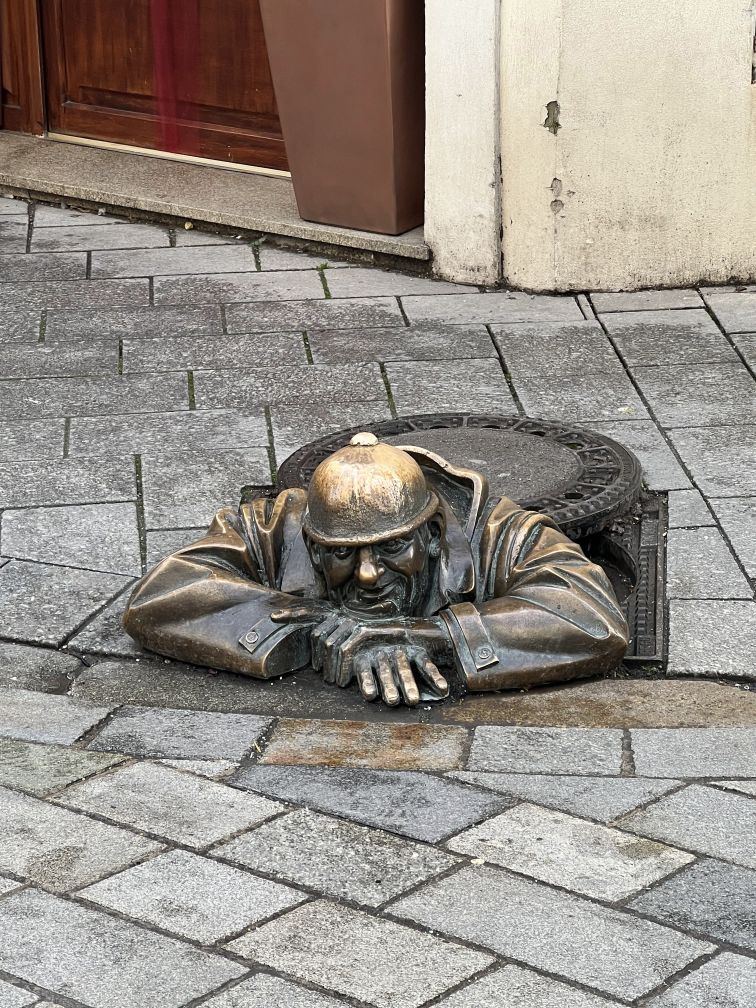 Bronze sculpture of a man in a manhole. His arms are cross and he is resting his chin on his arms. He is wearing a work helmet.