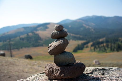 A cairn made of five dark stones stacked vertically upon a large boulder. In the background there are rolling green hills and large green mountains with an overcast sky above.