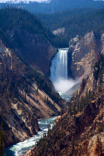 Evening view of Artist Point at the Grand Canyon of the Yellowstone. The rock walls of the canyon are various hues of reds and yellows, adorned with evergreen trees. There is a waterfall that appears white due to the heavy flow of the water. The river below is shades of blue white.