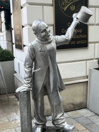 Silver and bronze sculpture of a smiling man. He has removed his tophat with his left hand in a greeting gesture.